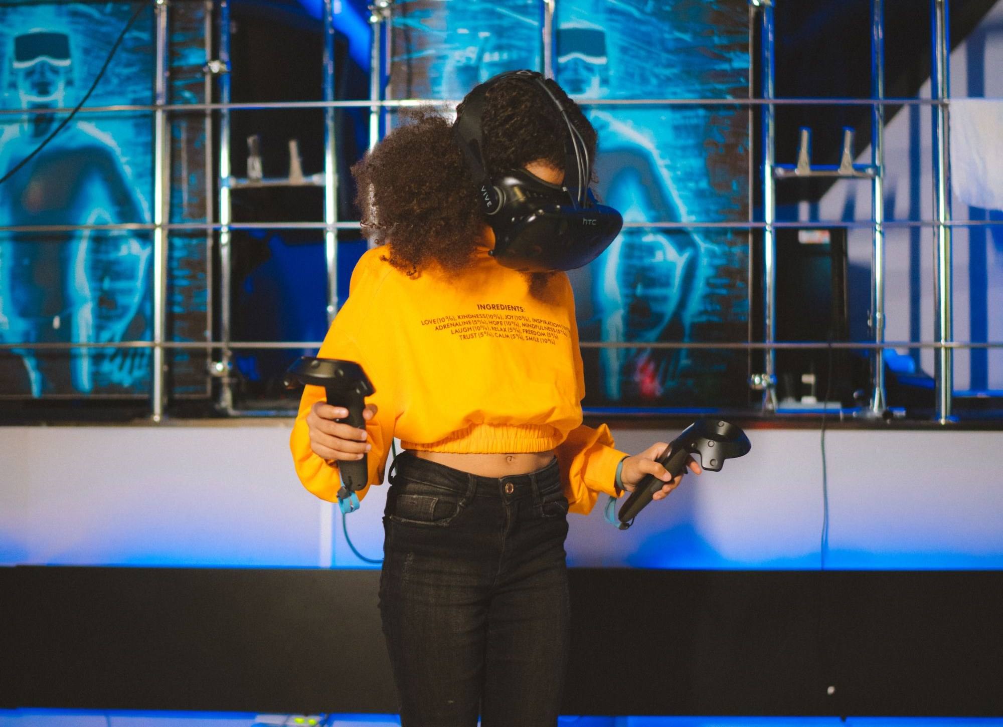 A girl in a yellow sweatshirt tries a virtual reality experience with two hand controllers.