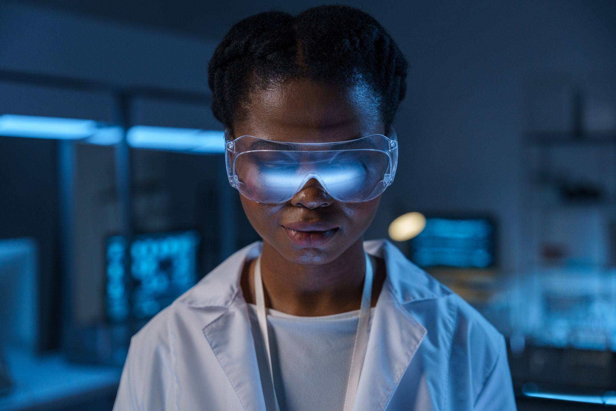 A young Black engineer wearing safety glasses in a laboratory lit with blue light.