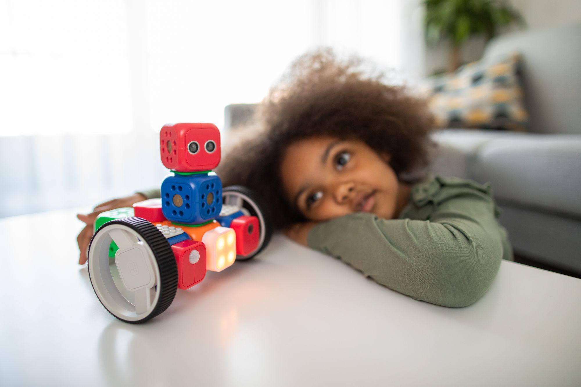 A young girl rests her head on a table beside a colourful toy robot.