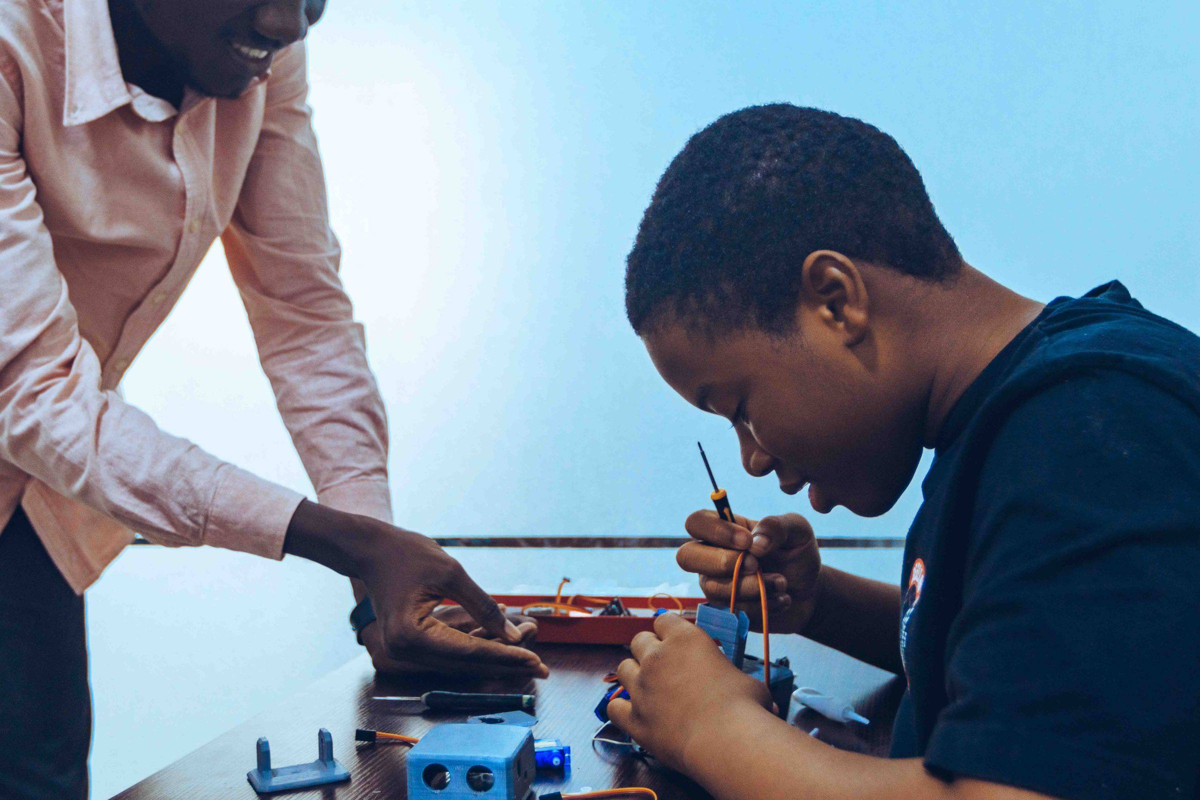 A boy concentrates as he wires a small robot arm while a mentor points to the next part, at a STEM workshop in Accra.