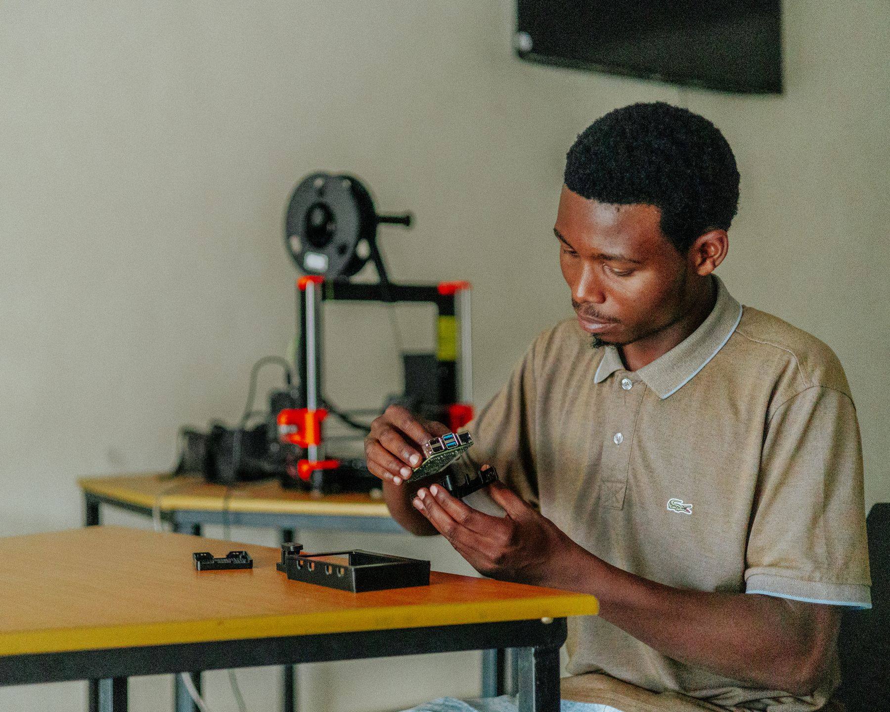 A young man assembles a small electronic part with a 3D printer on the bench behind him.